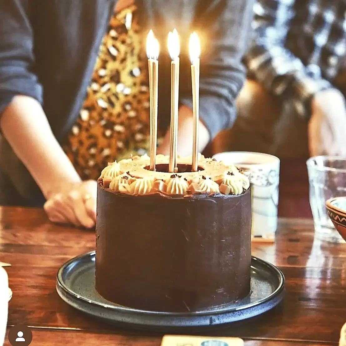 A chocolate birthday cake with 6 light candles sits upon a table surrounded by a group of people