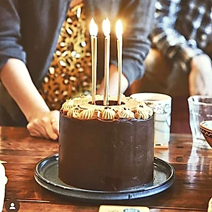 A chocolate birthday cake with 6 light candles sits upon a table surrounded by a group of people